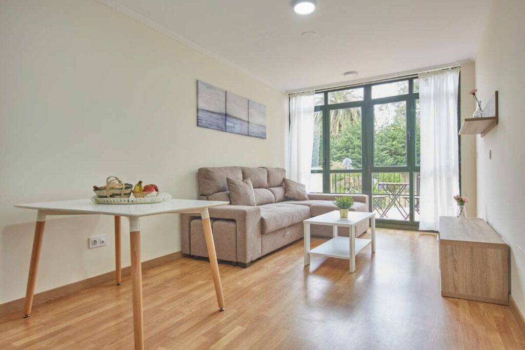 Salón de Apartagal con sofá beige, mesa de comedor y ventanal con acceso a terraza arbolada