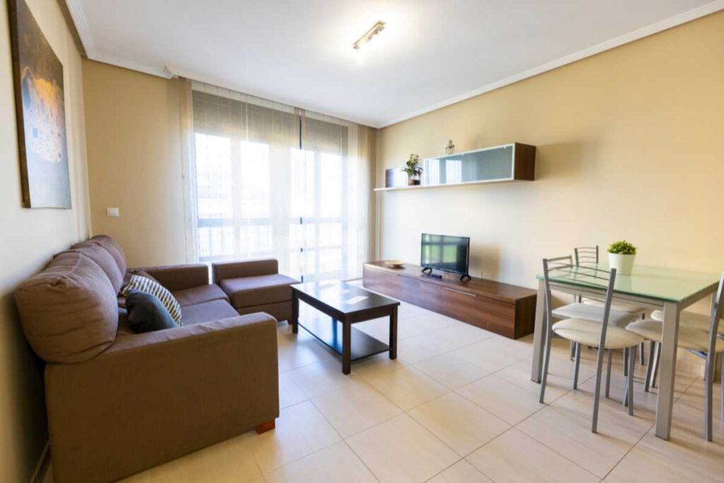 Salón comedor de Apartagal con sofá marrón, mesa de centro, mueble de TV y mesa con sillas junto a un ventanal luminoso