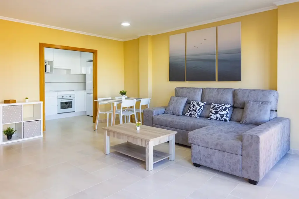 Salón con sofá gris, mesa de centro de madera y cocina blanca abierta al fondo en un apartamento de Apartagal