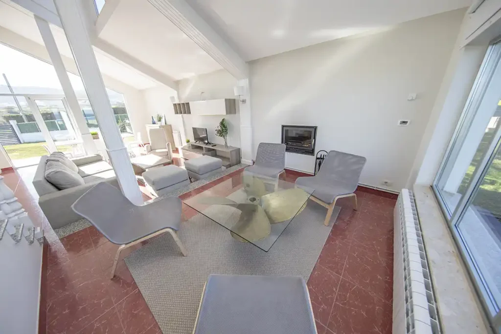 Salón amplio con techo a dos aguas, chimenea, sofá y butacas grises junto a ventanales con vistas al jardín