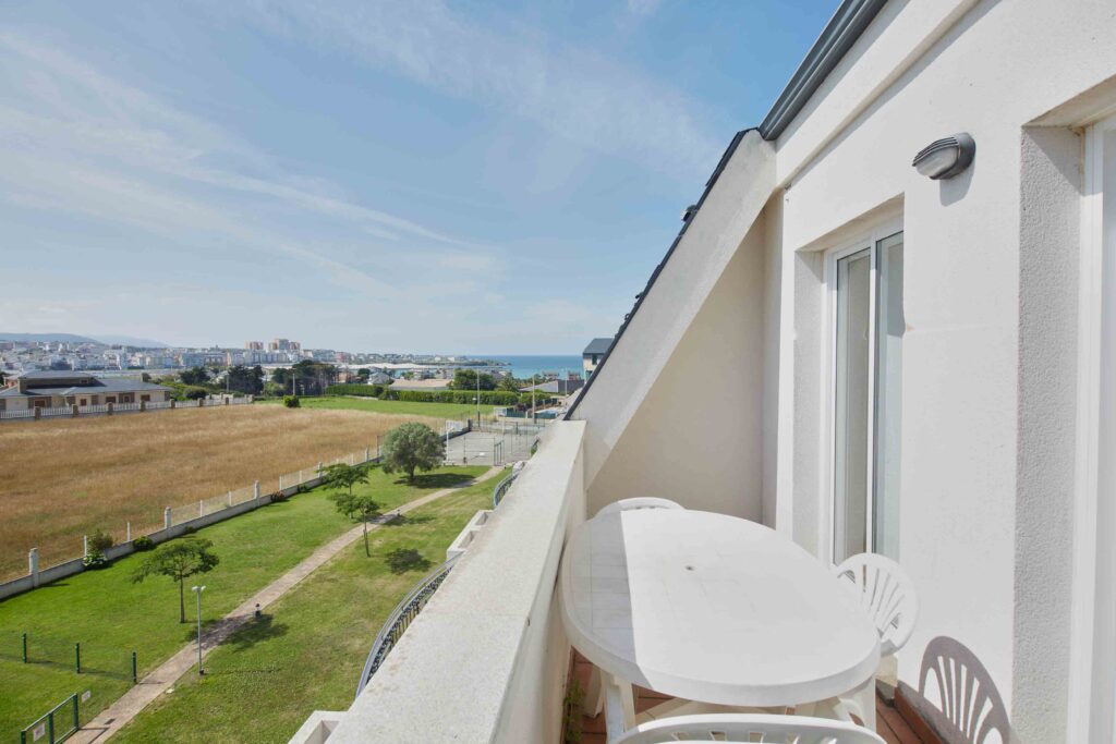 Terraza blanca con mesa y sillas junto a una fachada, con vistas a un parque, el pueblo y el mar al fondo