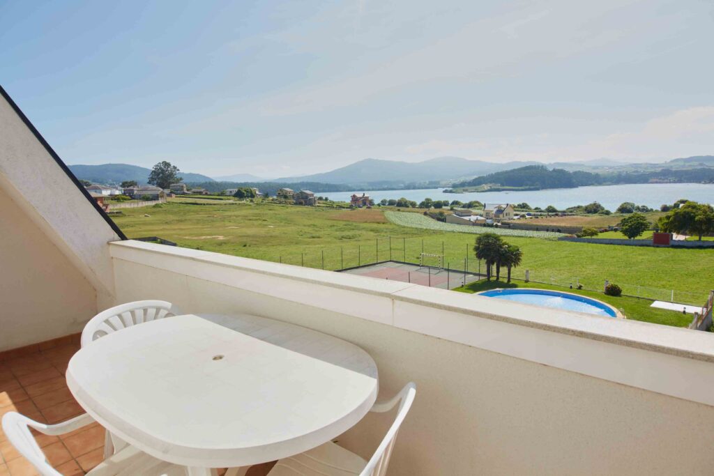 Terraza con mesa y sillas blancas, vistas a la ría, piscina, pista deportiva y montañas al fondo