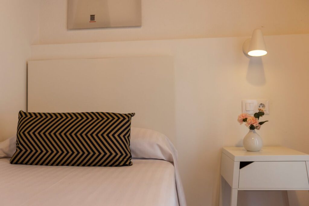 Dormitorio con cama blanca, cojín de rayas, mesita con jarrón de flores y lámpara de pared en la Casiña de Portochico