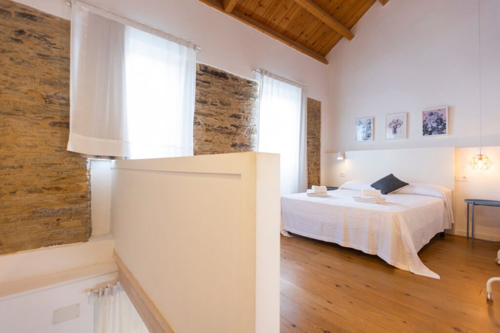 Dormitorio abuhardillado con cama de matrimonio, pared de piedra, vigas de madera y cortinas blancas en Apartagal