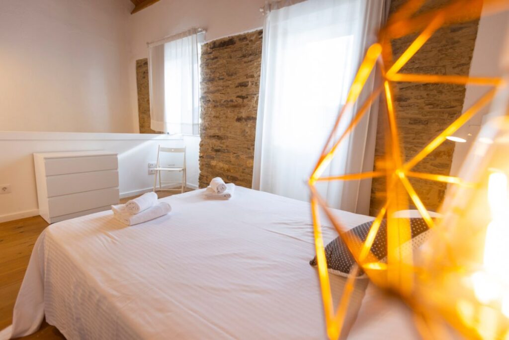Dormitorio con cama doble, toallas enrolladas, pared de piedra vista y cortinas blancas en un apartamento de Apartagal