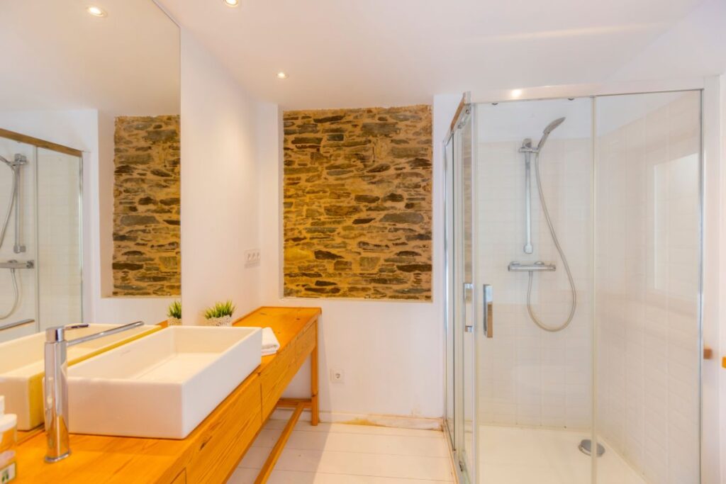 Baño con mueble de madera, dos lavabos, ducha con mampara de cristal y muro de piedra vista en Apartagal