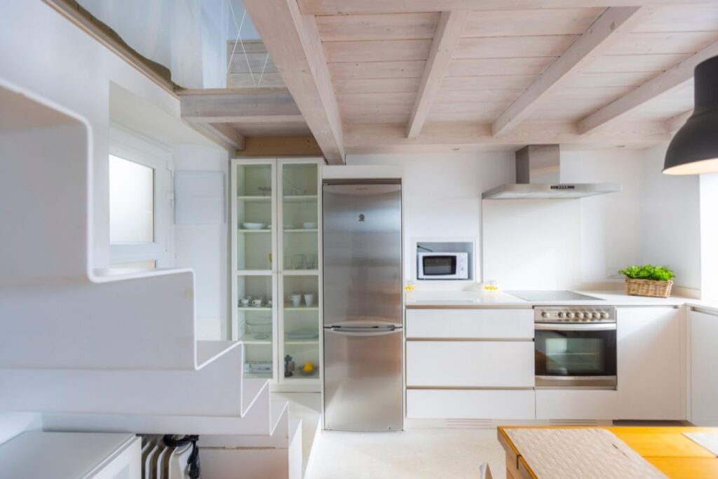 Cocina blanca a doble altura con vigas de madera, nevera de acero y escalera al altillo en Apartagal Foz