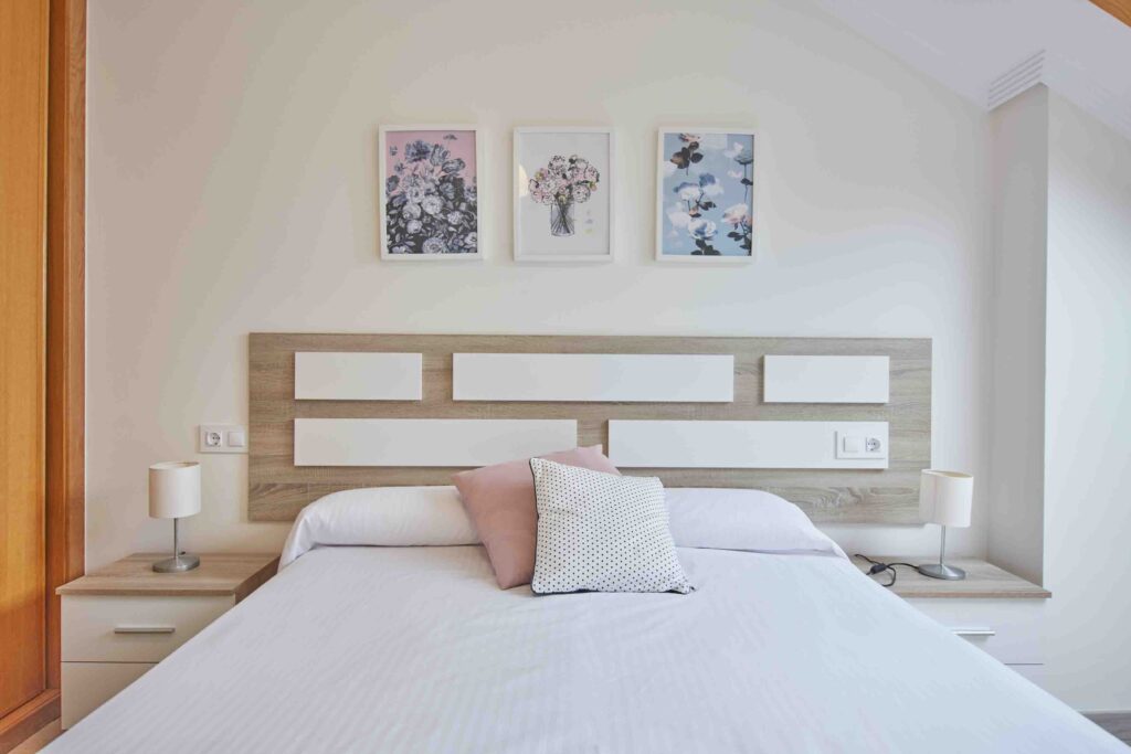 Dormitorio de matrimonio con cabecero de madera, cojín rosa y tres láminas florales sobre la cama