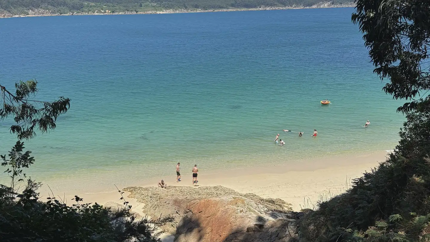 Vista elevada de una playa de O Vicedo con arena, agua turquesa y varias personas bañándose, rodeada de monte